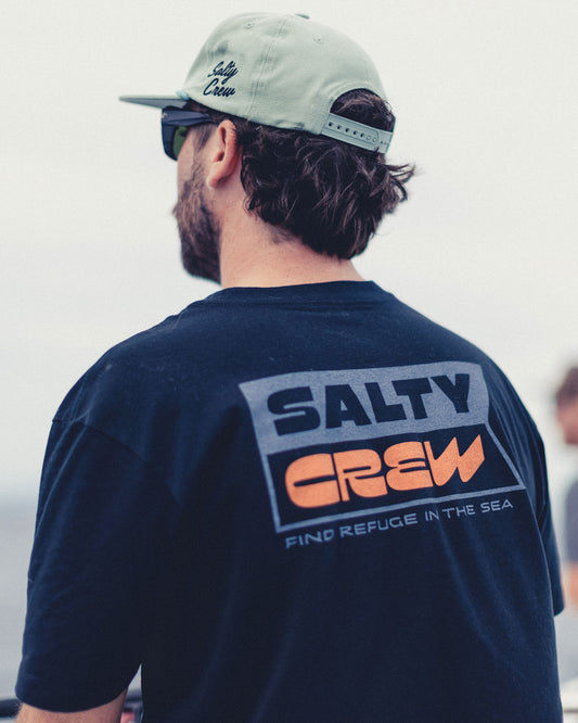 A man wearing a black Inverse Flag Crew Tee, a 100% cotton short-sleeved shirt with a screenprinted rectangular graphic on the back that reads SALTY CREW in large text and FIND REFUGE IN THE SEA below it.