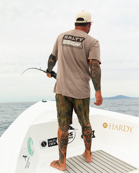 A tattooed man in a Salty Crew Inverse Flag Crew Tee - Cinder and camouflage shorts stands barefoot on a boat, holding a fishing rod as he faces the ocean, with distant land visible beneath a cloudy sky.