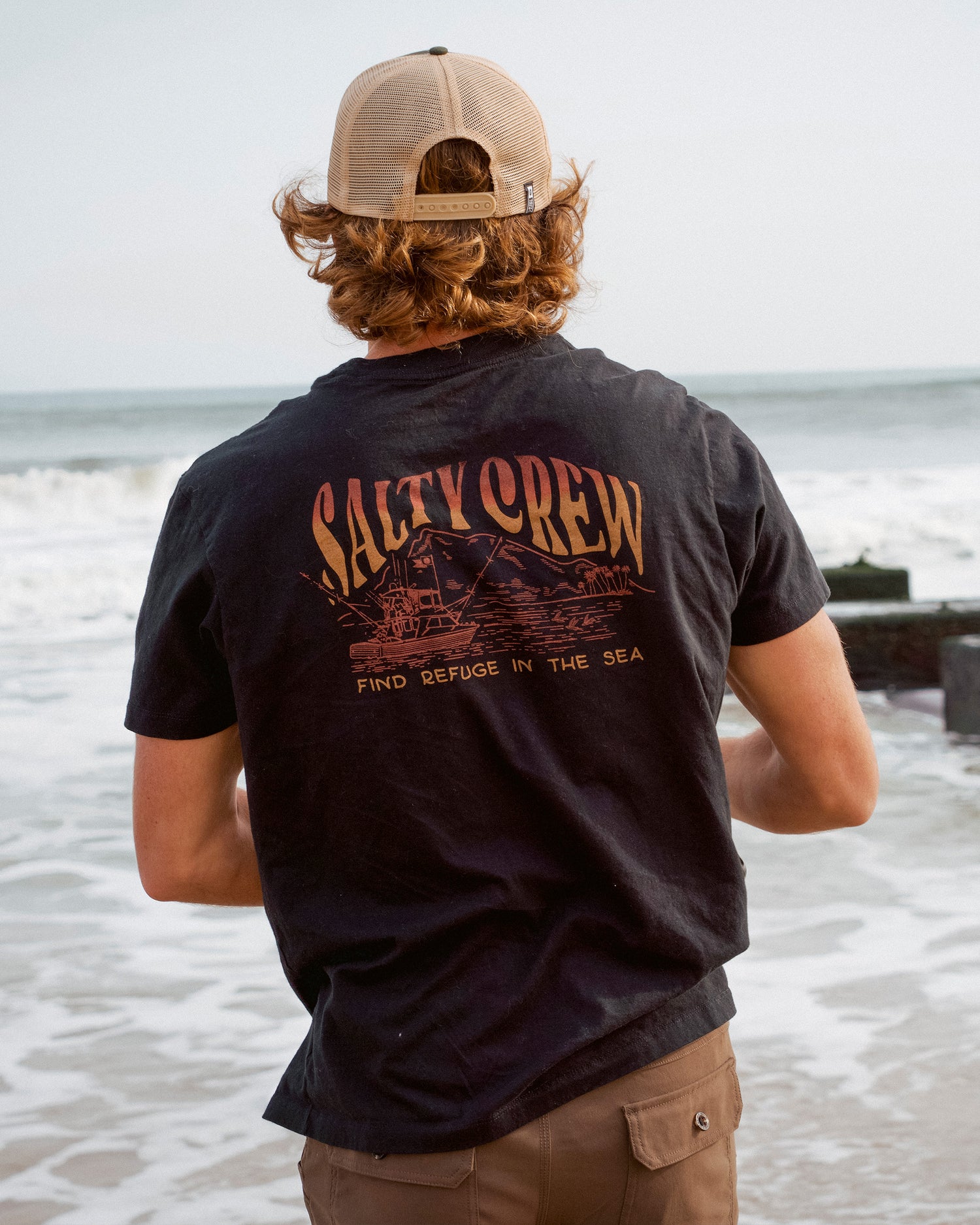 A person with wavy hair, wearing a black Island Crew Tee from Salty Crew and a cap, stands facing the ocean. The combed ringspun cotton shirt features a screenprinted graphic of a boat along with the words “Find Refuge in the Sea.”.