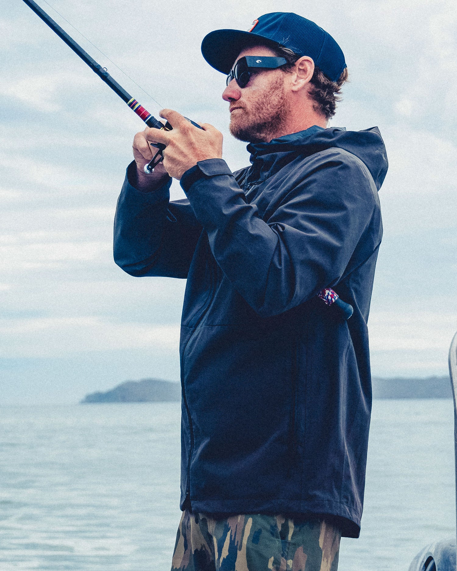 A man in a durable, black Salty Crew Long Range Jacket and camouflage shorts fishes from a boat.