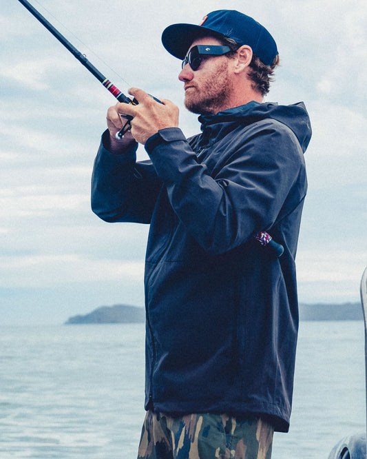 A man in a durable, black Salty Crew Long Range Jacket and camouflage shorts fishes from a boat.