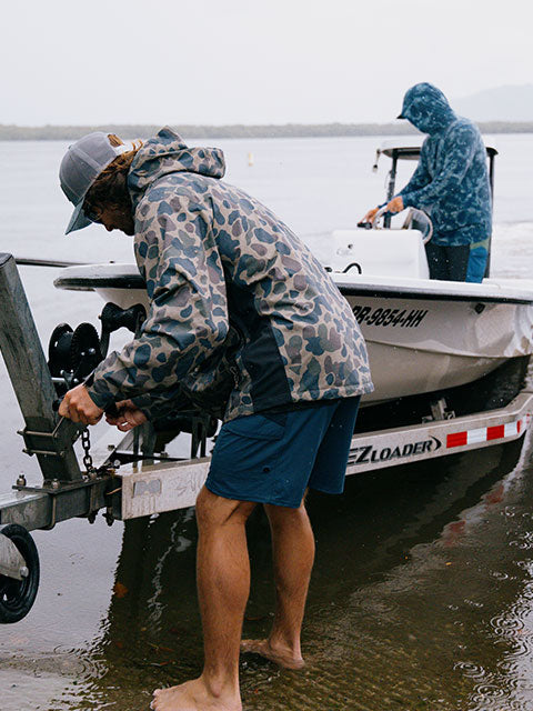Wearing Salty Crew’s Long Range Jacket in Sand Camo, two people prepare a boat for launch at the water's edge; one adjusts the trailer while the other stands ready in the boat. Clad in this durable outerwear, they brace against the cool, overcast day, ready for adventure on the water.