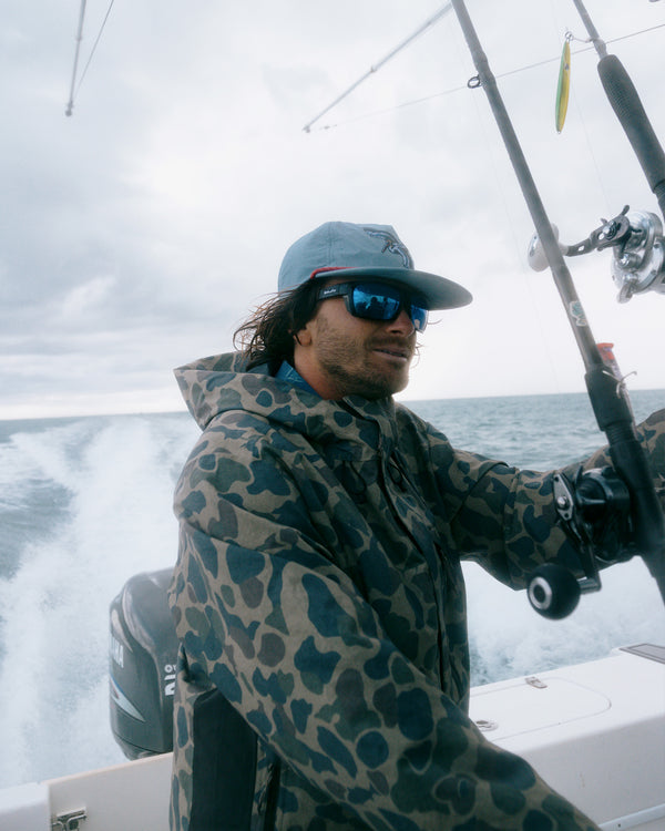 A person wearing the Salty Crew Long Range Jacket in Sand Camo—with 20K breathability—along with sunglasses and a cap, steers a boat on the ocean while holding fishing rods. The water and cloudy sky are visible in the background.