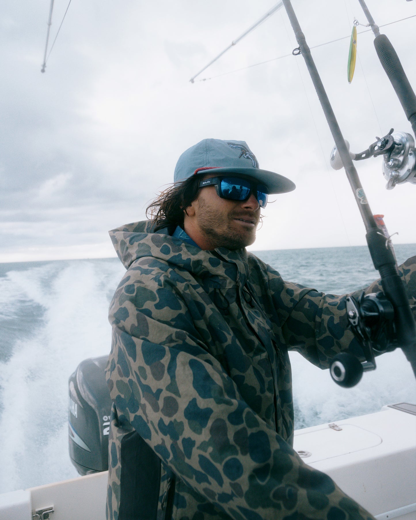 A person wearing the Salty Crew Long Range Jacket in Sand Camo—with 20K breathability—along with sunglasses and a cap, steers a boat on the ocean while holding fishing rods. The water and cloudy sky are visible in the background.