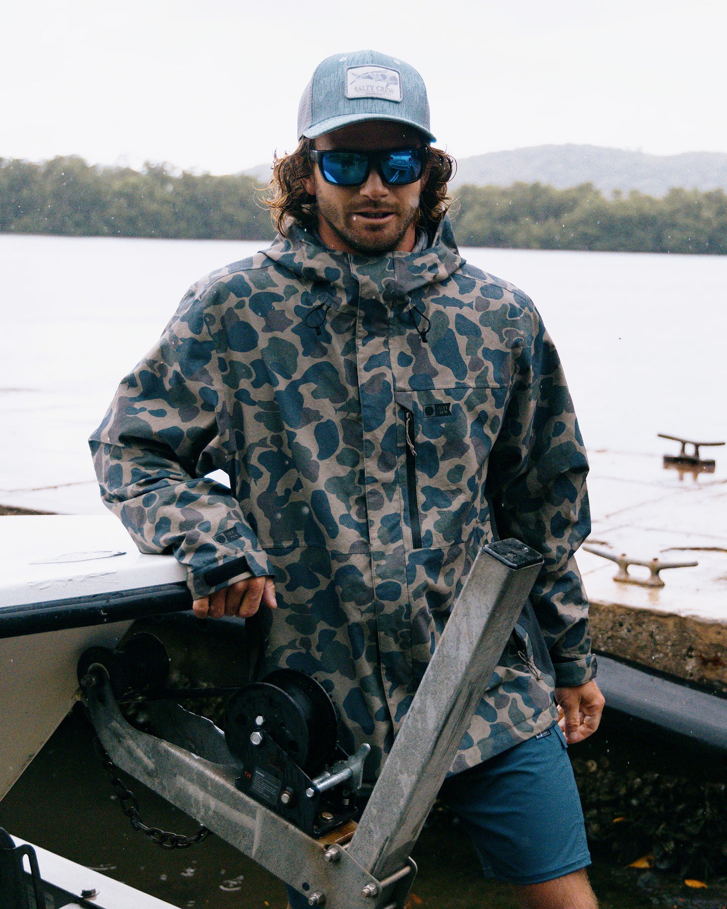 A man wearing the Salty Crew Long Range Jacket in Sand Camo, along with a blue hat and sunglasses, stands by a boat near the water, with trees and hills visible in the background on a cloudy day.