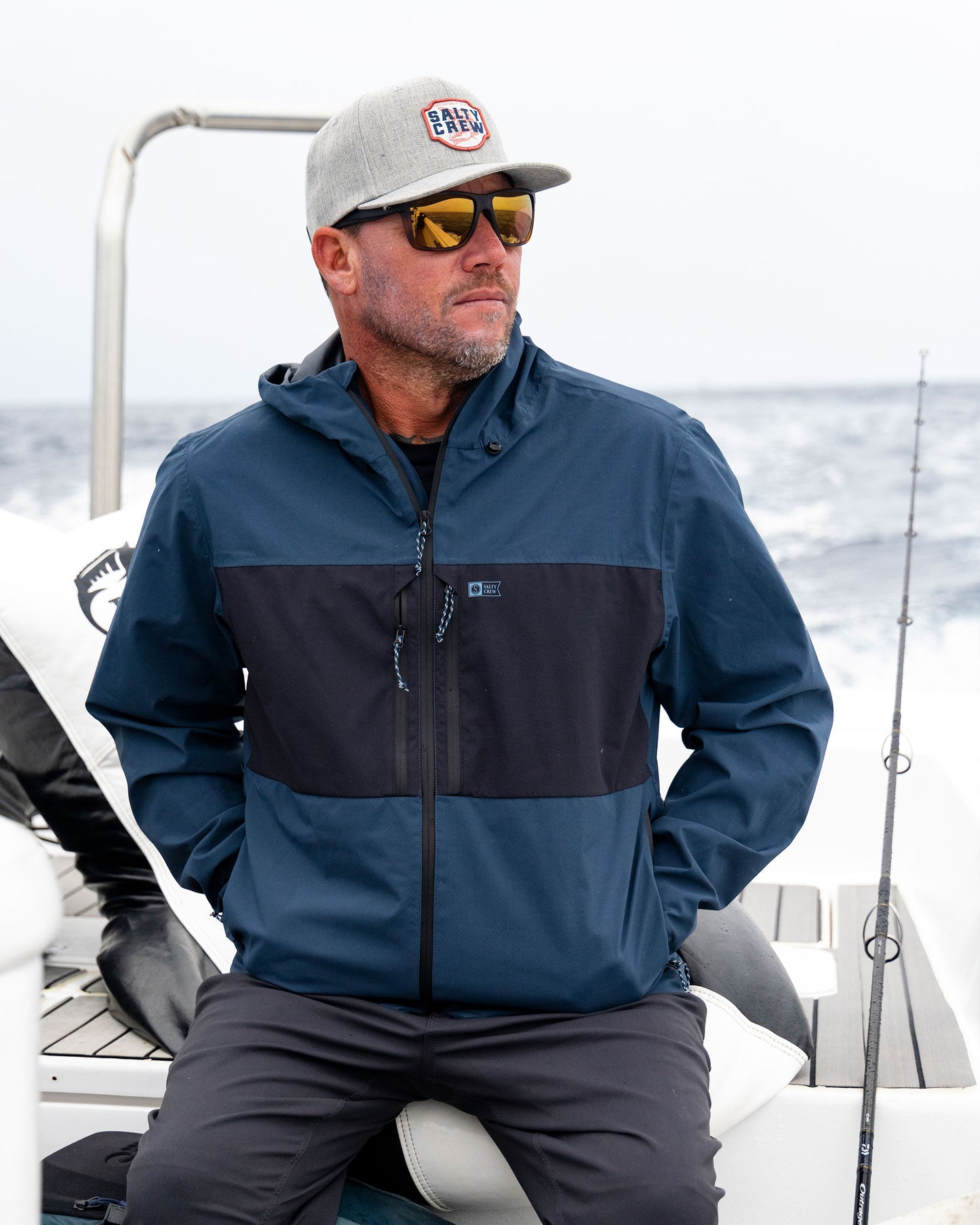 A man wearing a navy Salty Crew Long Range Jacket, a baseball cap, and sunglasses sits on the deck of a boat with a fishing rod nearby.
