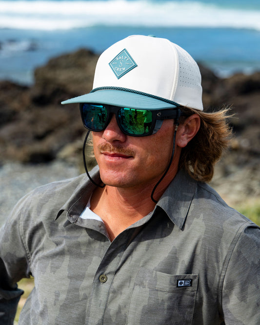 A person wearing an off-white and ocean Long Range Tech Snapback with a diamond-shaped logo and mirrored sunglasses outdoors.