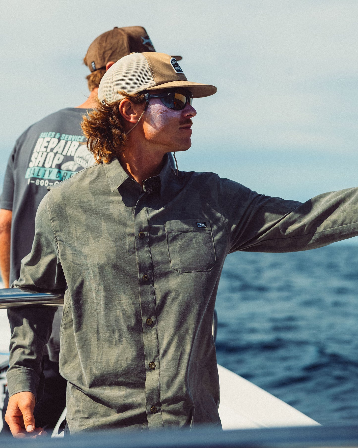 Man wearing a long-sleeved, moisture-wicking olive green camouflage Drifter Long Sleeve UV Button Up - Ranger, sunglasses, and a cap while standing on a boat.
