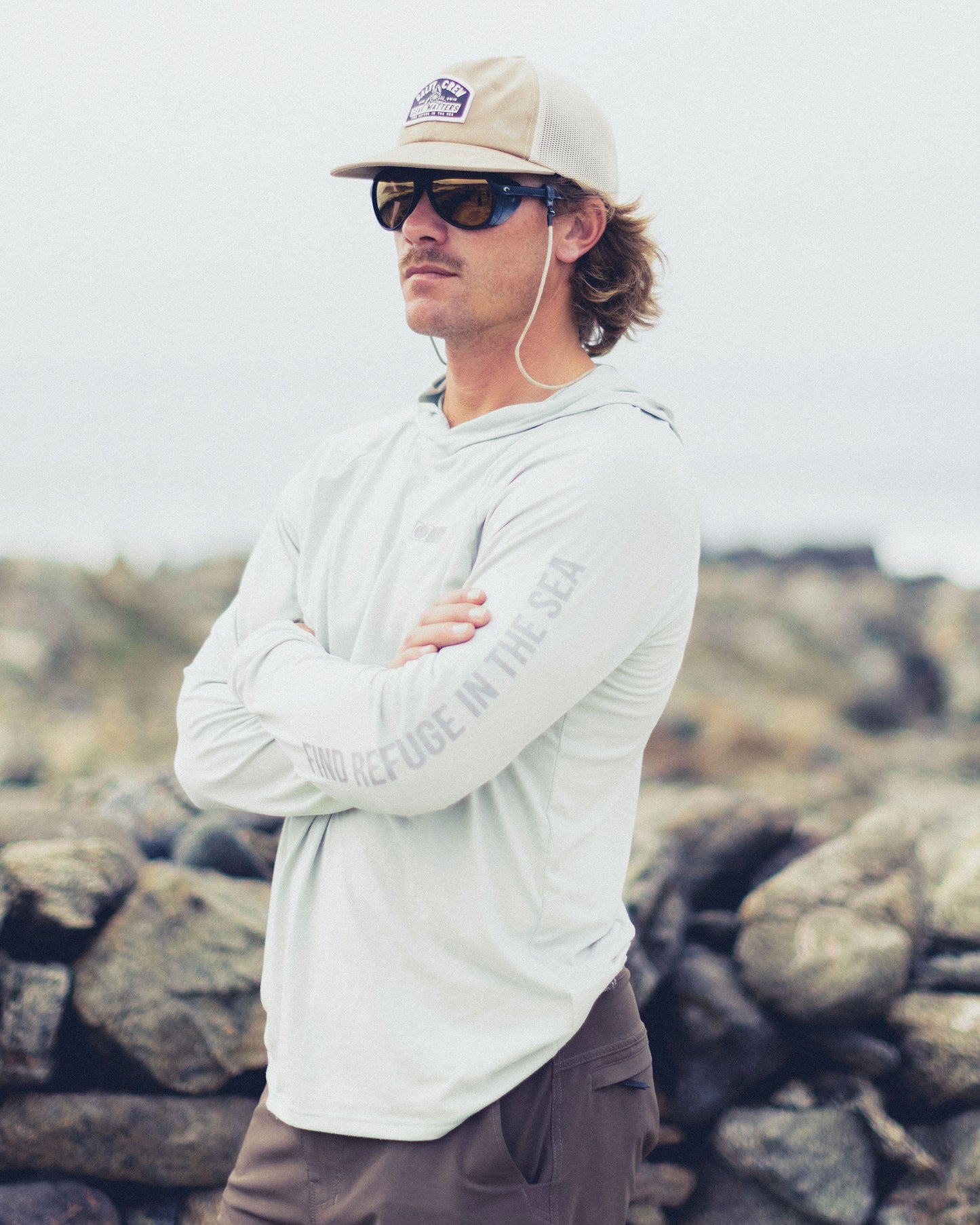 A man wearing a light grey Mariner Lite UV Hood - Aqua Grey with FIND REFUGE IN THE SEA on the sleeve, sunglasses, and a hat standing in front of rocky terrain.