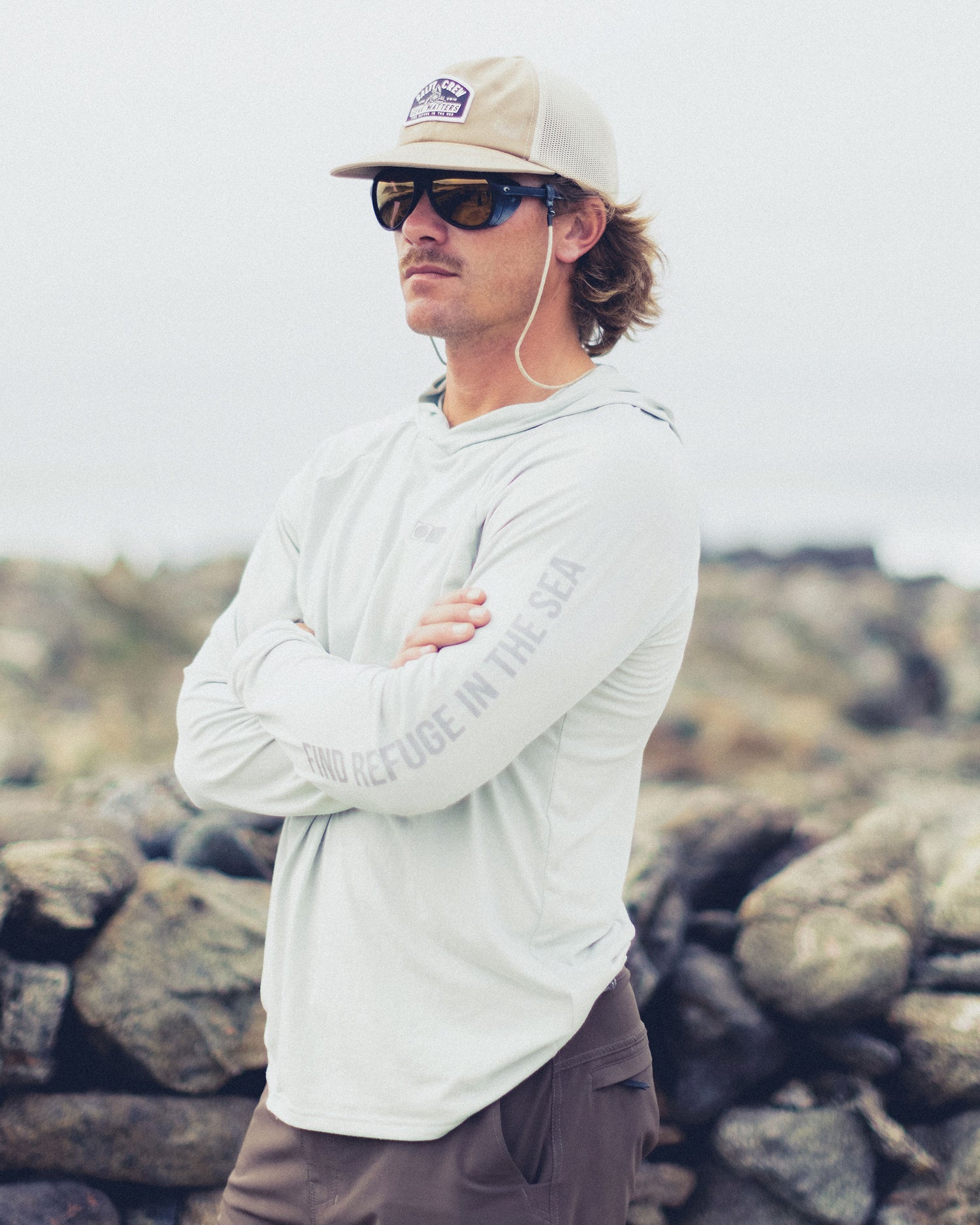 A man wearing a light grey Mariner Lite UV Hood - Aqua Grey with FIND REFUGE IN THE SEA on the sleeve, sunglasses, and a hat standing in front of rocky terrain.