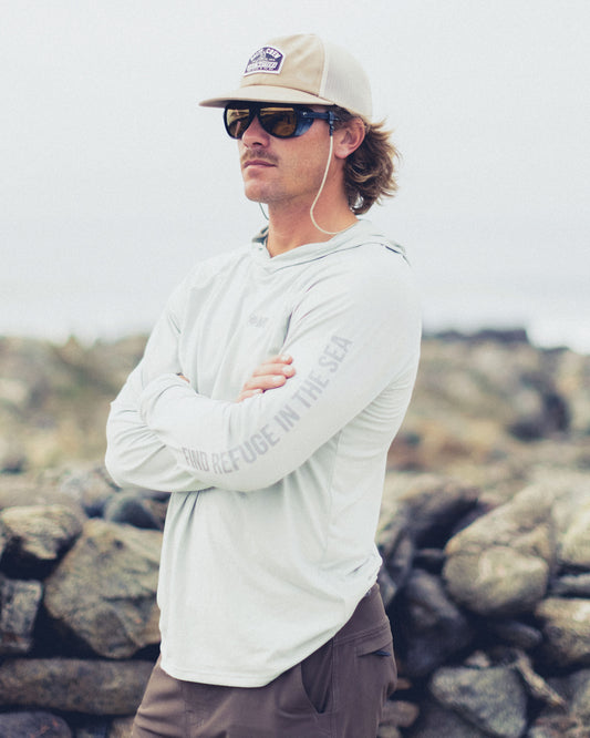 A man wearing a light grey Mariner Lite UV Hood - Aqua Grey with FIND REFUGE IN THE SEA on the sleeve, sunglasses, and a hat standing in front of rocky terrain.