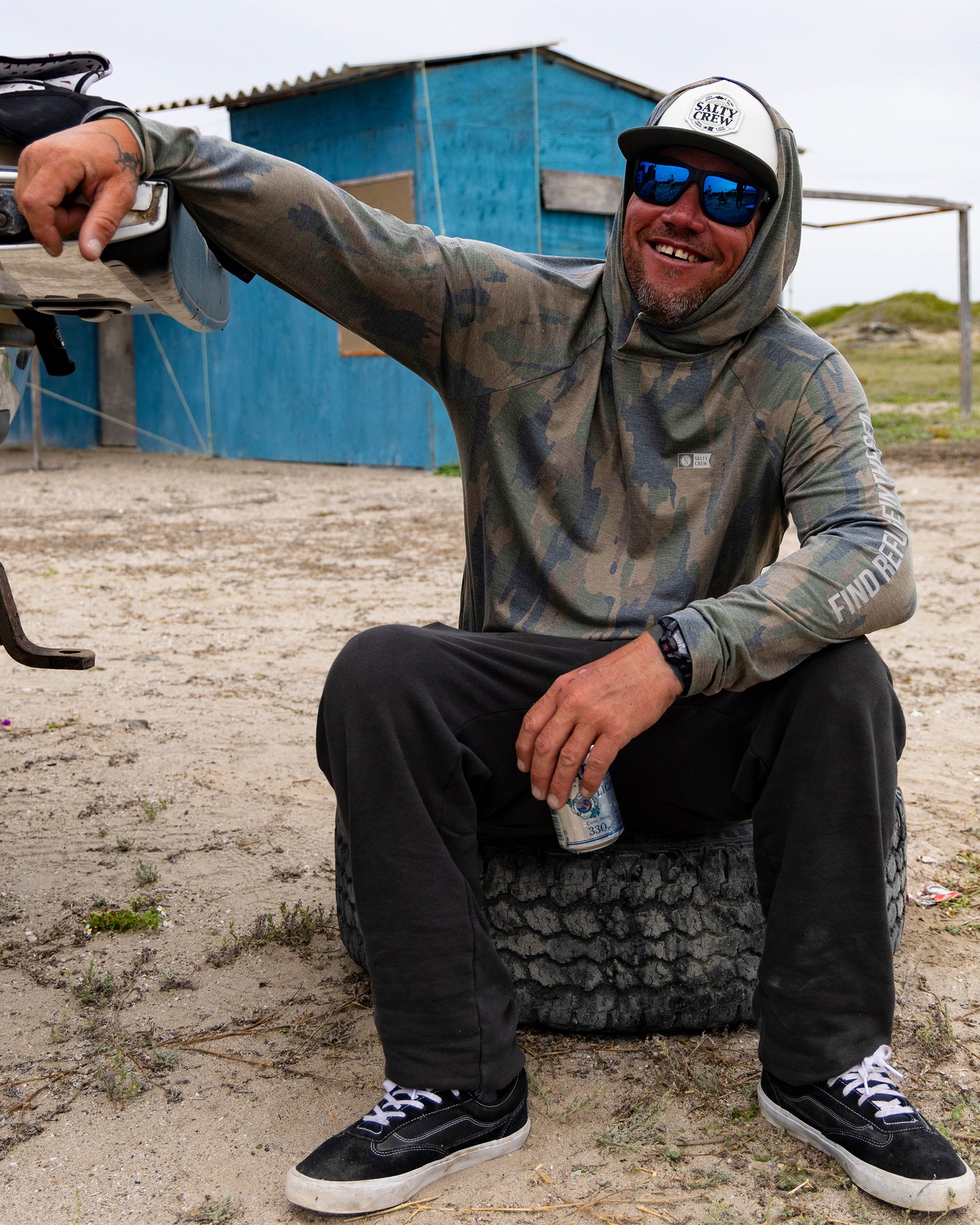 A man in sunglasses and the Salty Crew Mariner Lite UV Hood - Ranger, featuring UPF 50+ sun protection, sits on a tire, smiling and holding a can. He gestures with one hand. A blue wooden building stands in the background on a sandy surface.
