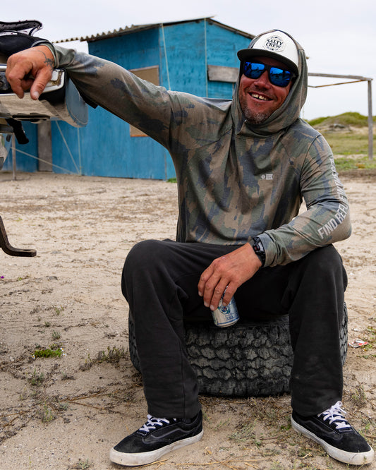 A man in sunglasses and the Salty Crew Mariner Lite UV Hood - Ranger, featuring UPF 50+ sun protection, sits on a tire, smiling and holding a can. He gestures with one hand. A blue wooden building stands in the background on a sandy surface.