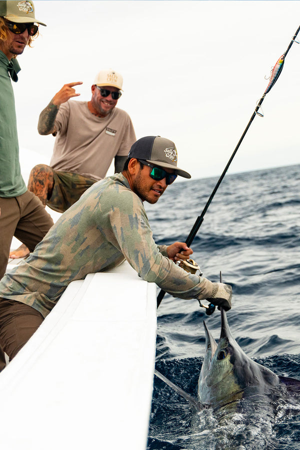Three men fish at sea from a boat; one holds a fishing rod with a large marlin at the water’s surface, while another flashes a peace sign and smiles. Outfitted in Salty Crew’s Mariner Lite UV Hood - Ranger shirts that wick away moisture, they stay comfortable under the sun as they enjoy their adventure, with the sky and ocean as their backdrop.