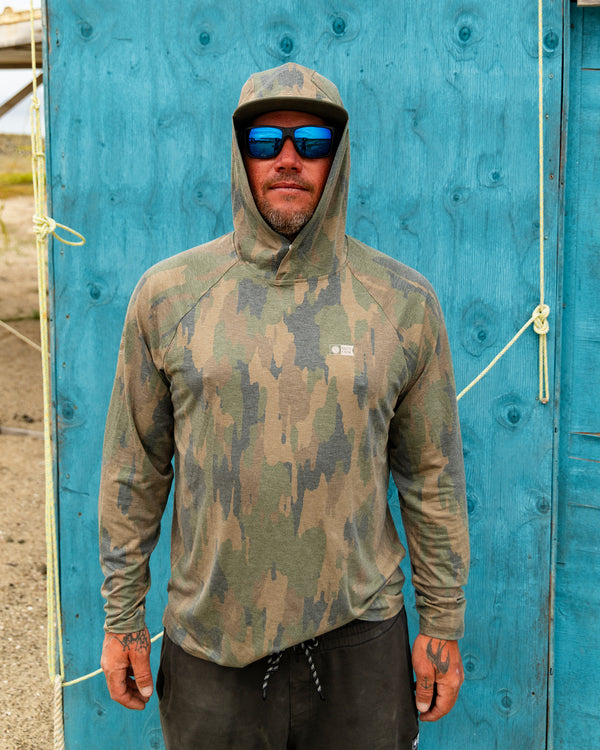 A man wearing the Salty Crew Mariner Lite UV Hood - Ranger, along with sunglasses and a cap, stands in front of a bright blue wooden wall adorned with rope. Displaying tattoos on his hands, he looks directly at the camera.