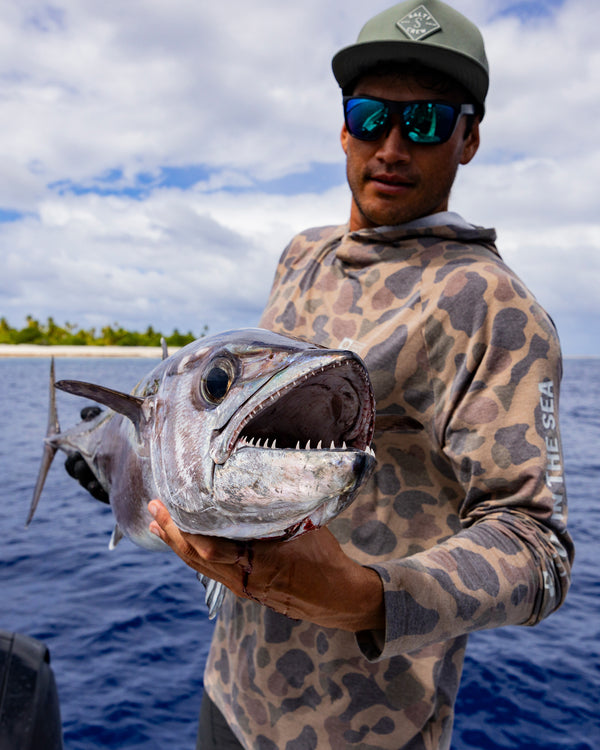 A man in sunglasses and a Salty Crew Mariner Lite UV Hood - Tan Camo holds a large, sharp-toothed fish over the water, with a tropical shoreline and partly cloudy sky in the background.