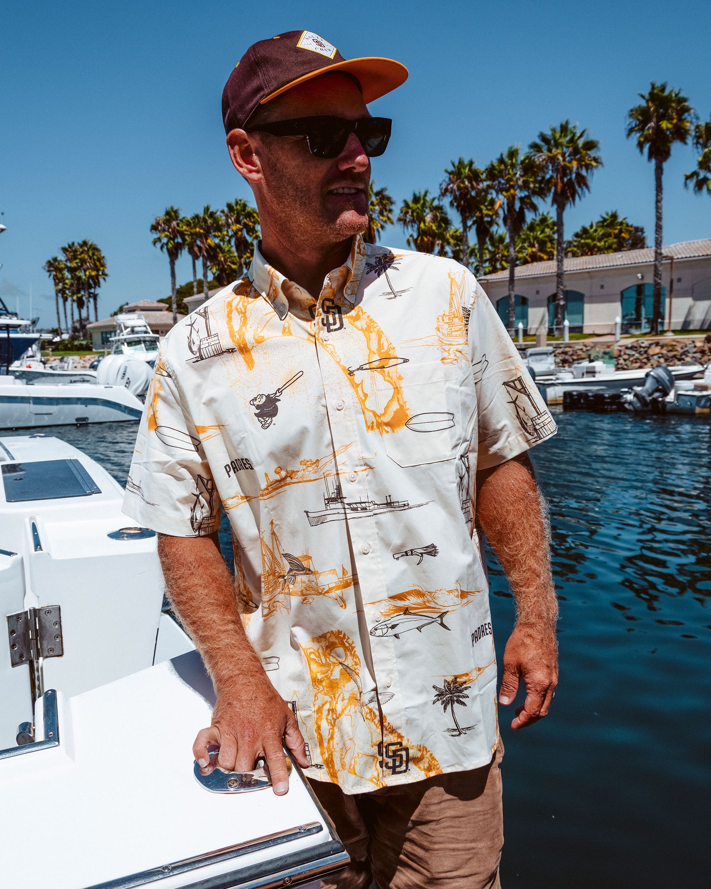 A man in sunglasses, a cap, and the Salty Crew Padres x Reyn Spooner UV Button Up - Bone short-sleeve shirt stands by a marina with boats, palm trees, and buildings in the background on a sunny day.