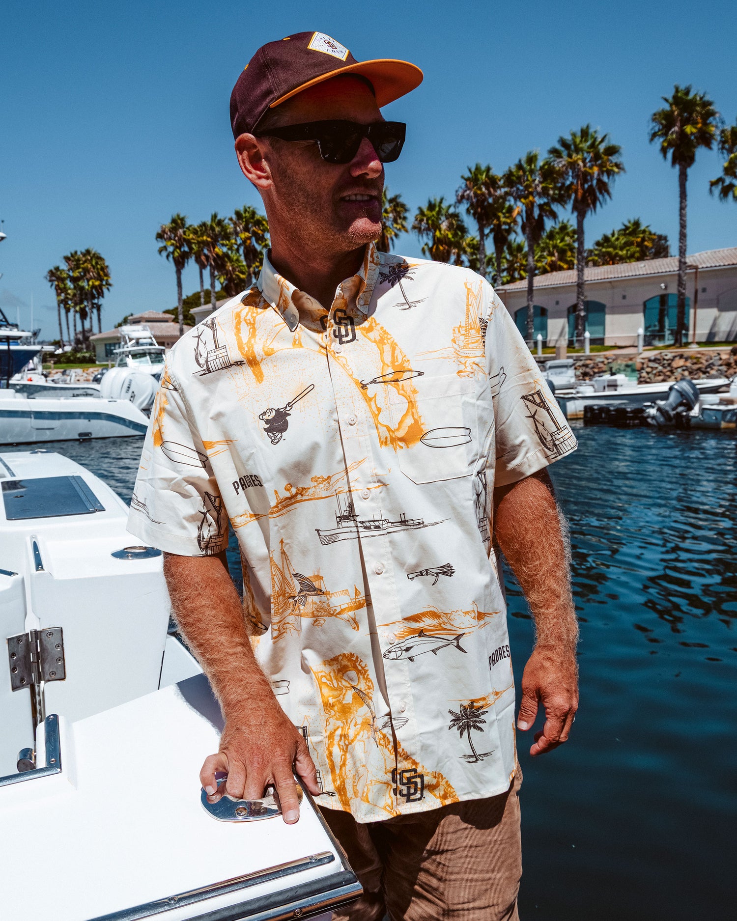 A man in sunglasses, a cap, and the Salty Crew Padres x Reyn Spooner UV Button Up - Bone short-sleeve shirt stands by a marina with boats, palm trees, and buildings in the background on a sunny day.
