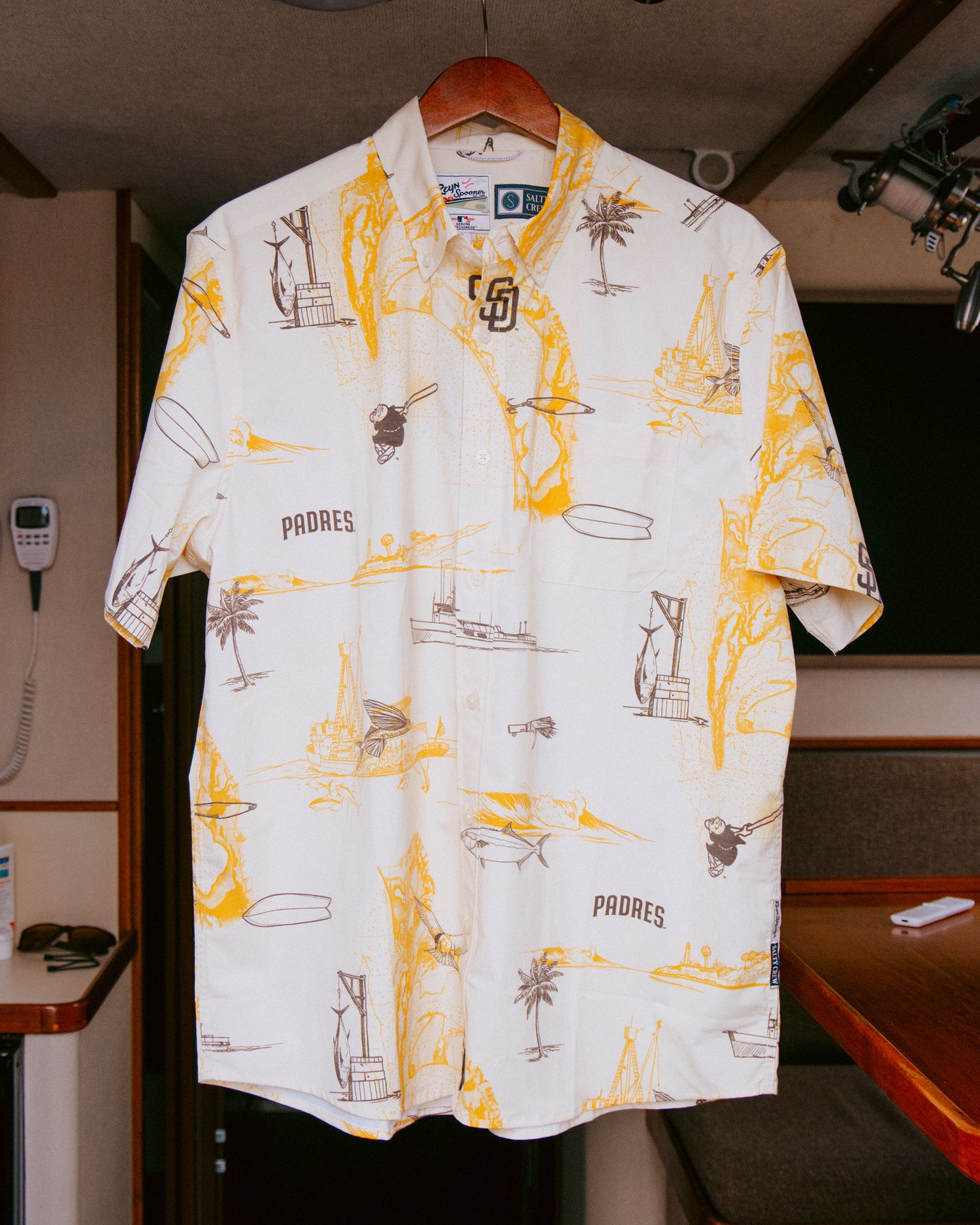 Padres x Reyn Spooner UV Button Up - Bone by Salty Crew, a short-sleeve bone-colored button-up shirt featuring yellow and brown nautical and Padres-themed illustrations like palm trees, boats, and the word “PADRES,” designed in collaboration with Reyn Spooner for the San Diego Padres, pictured hanging inside a room on a boat.