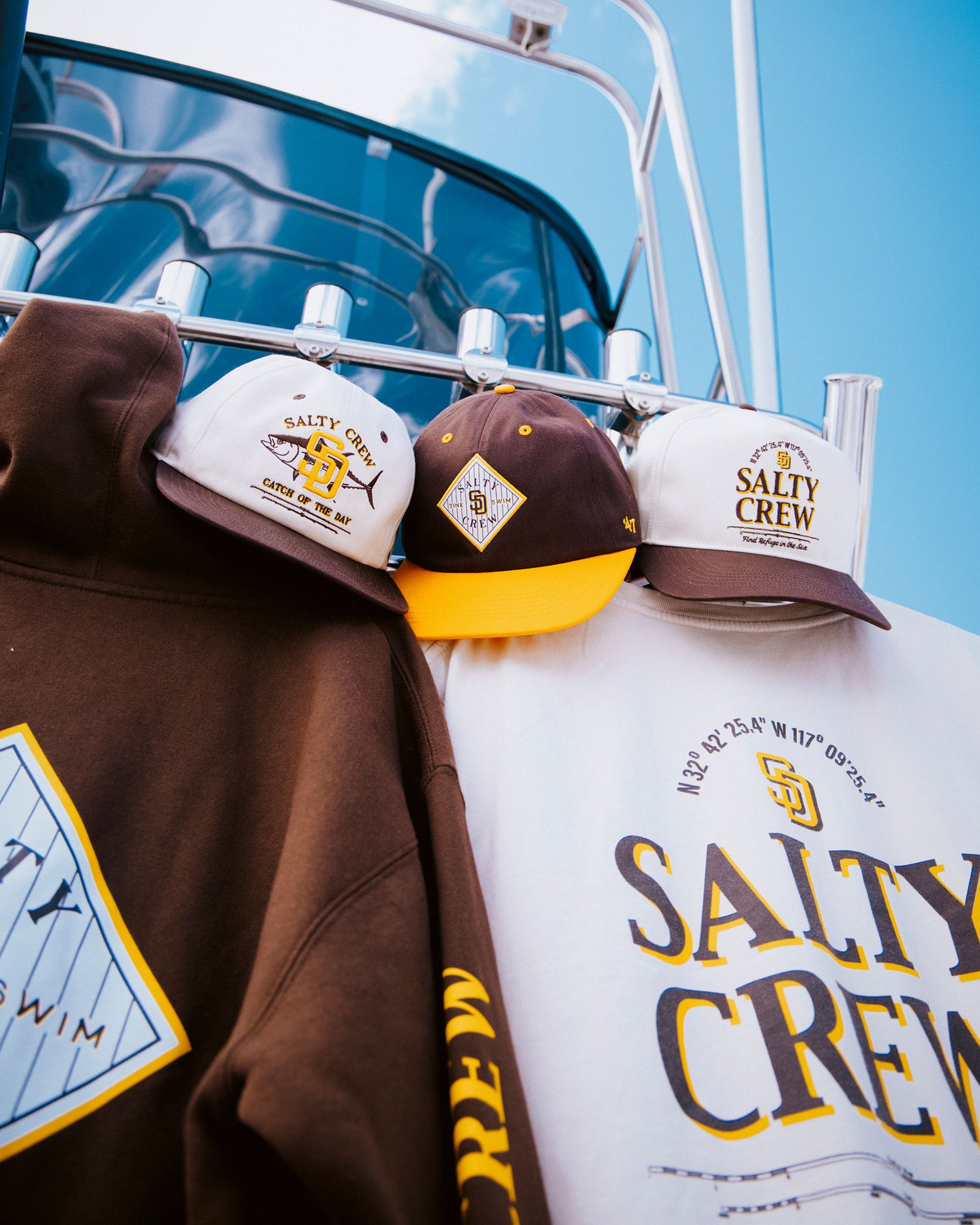 Three Salty Crew hats in brown, white, and yellow are displayed on a metal rack beside the Padres Tippet Fleece - Bone hoodie from Salty Crew and a white shirt, both featuring the brand’s iconic logos. This limited edition collection stands out against the backdrop of a blue boat, offering exclusive style for fans and adventurers alike.