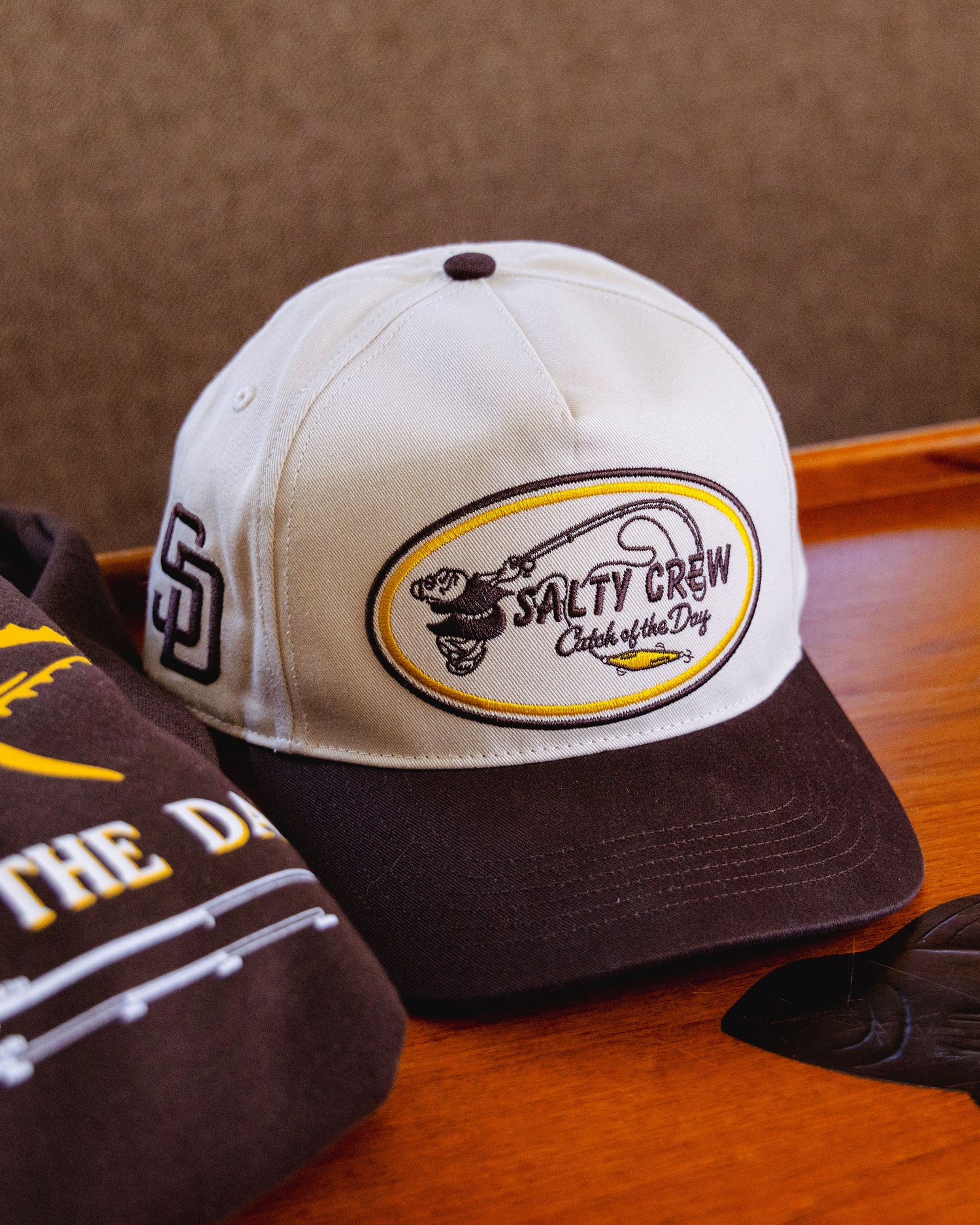 A limited edition Salty Crew Padres Swingin Friar Hat in bone, featuring a unique patch and fish graphic, rests on a wooden surface beside a brown garment—delivering standout style to rival your favorite ’47 Brand or San Diego Padres hat.