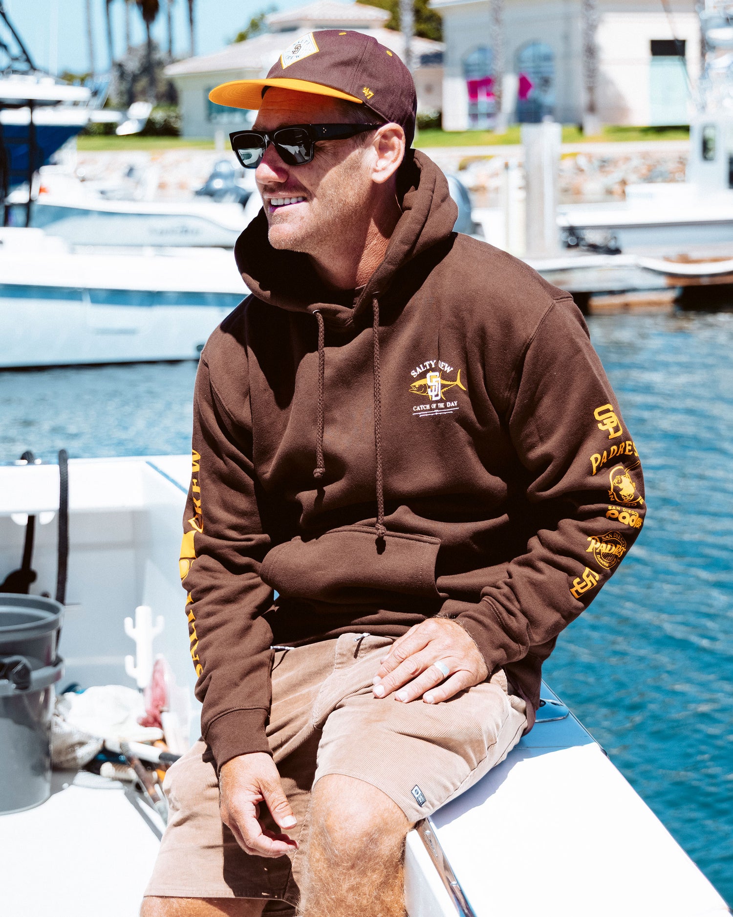 A man wearing the Salty Crew Padres Catch of the Day Fleece in brown, along with a matching cap and sunglasses, sits on a white boat docked in a marina, smiling on a sunny day.