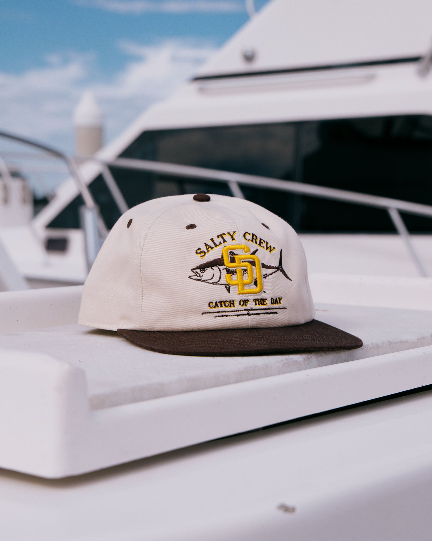 A Salty Crew Padres Catch of the Day Hat in bone rests on a white surface. In the background, a blurred yacht and blue sky can be seen.