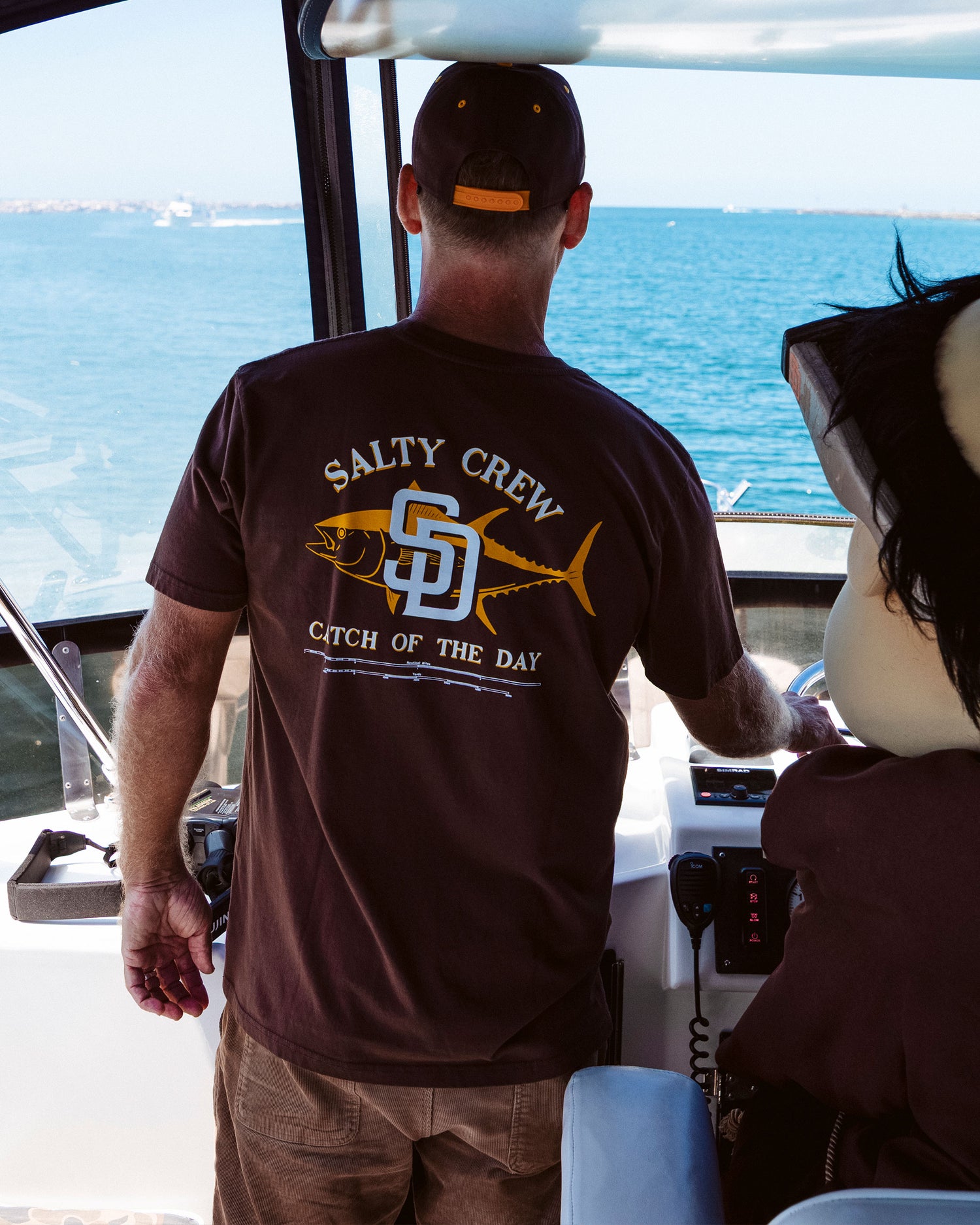 Wearing the Salty Crew Padres Catch of the Day Tee in brown, a person stands at the helm of a boat, steering on a clear day with calm blue water in the background.