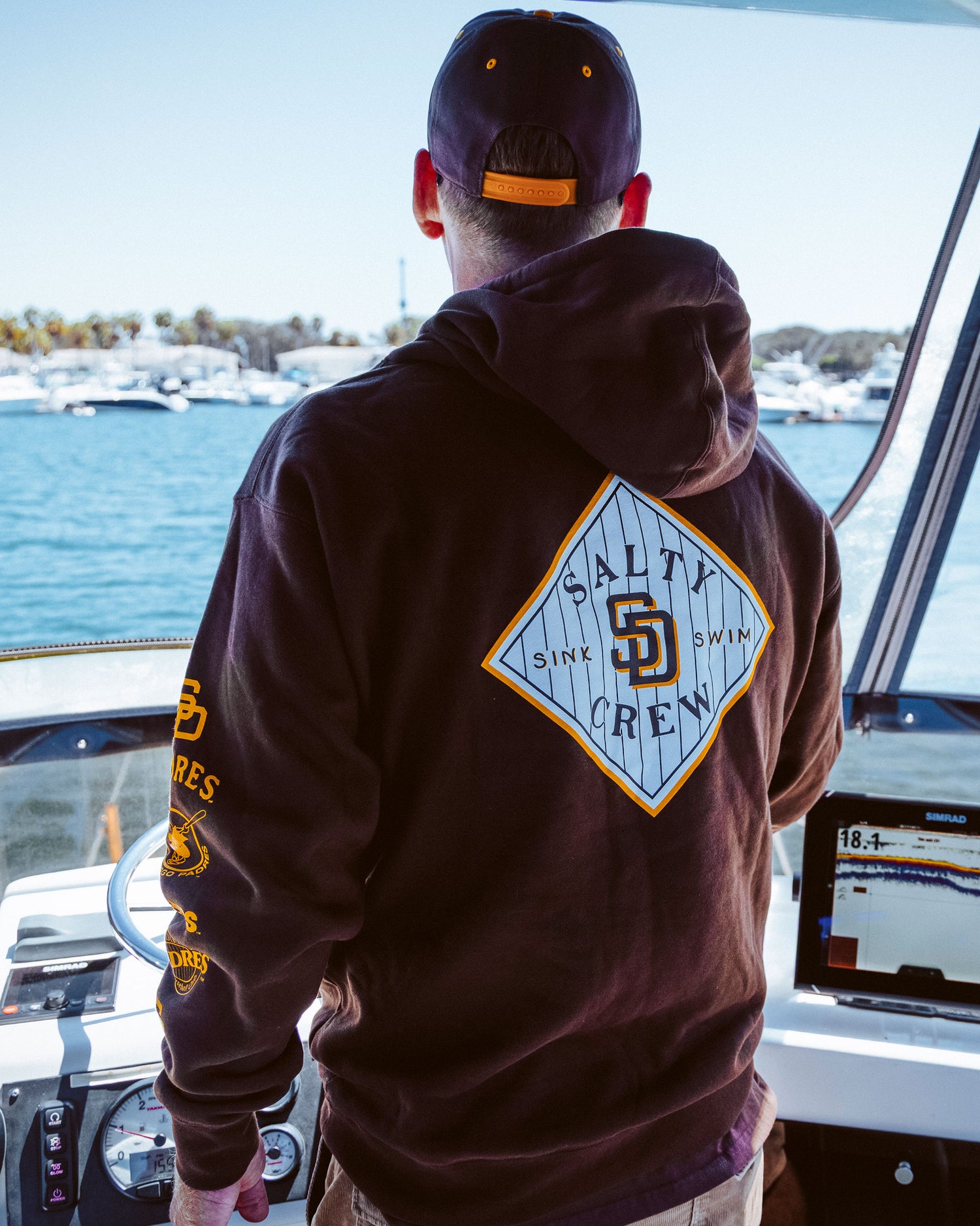 A person wearing a Salty Crew Padres Tippet Fleece – Brown hoodie and cap steers a boat on a sunny day, with boats and trees visible in the background near the water.