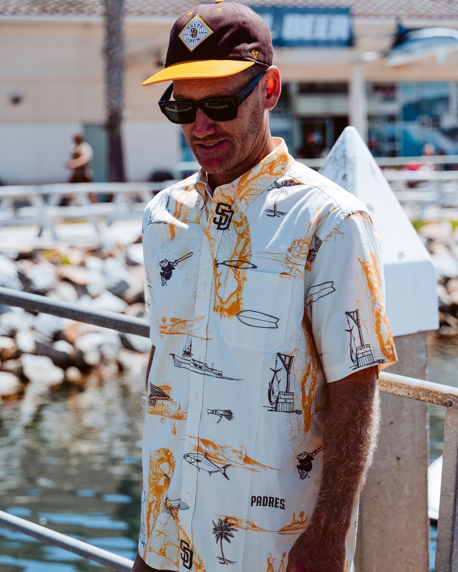 A man wearing sunglasses, a Salty Crew Padres Tippet Hat in brown and gold, and a patterned white button-up shirt stands by a waterfront railing on a sunny day.