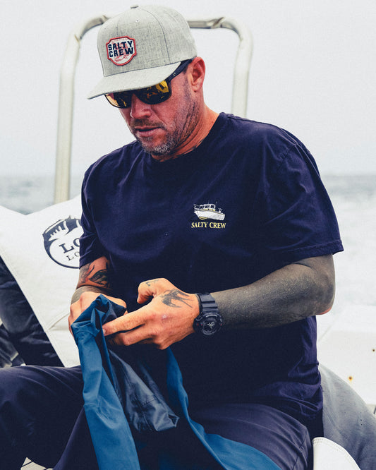 A man wearing a grey Salty Crew Red Fish Snapback - Heather Grey and a dark navy t-shirt holds a blue garment while sitting on a boat.