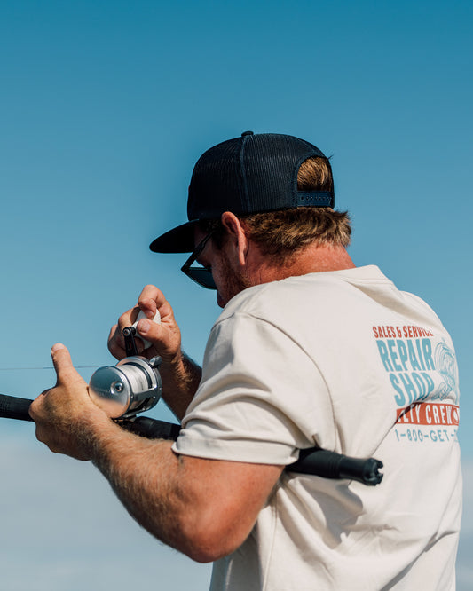 A man in a cap and sunglasses adjusts a fishing reel on a rod. He wears the Salty Crew Reel Repair Crew Tee in Bone—a lightweight, screenprinted combed ringspun cotton t-shirt featuring a repair shop logo—standing against a clear blue sky.