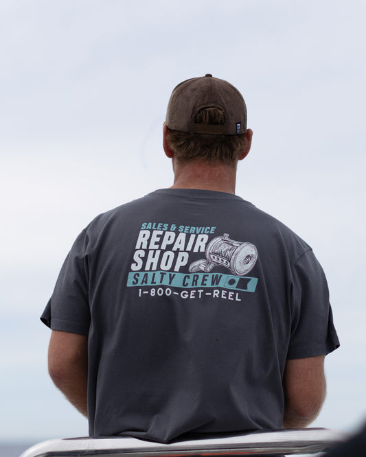 A person wearing the Salty Crew Reel Repair Crew Tee in dark grey and a brown cap faces away, displaying the shirt’s screenprinted “Sales & Service Repair Shop, Salty Crew, 1-800-GET-REEL” text with a reel graphic. The sky and part of a boat are visible in the background.