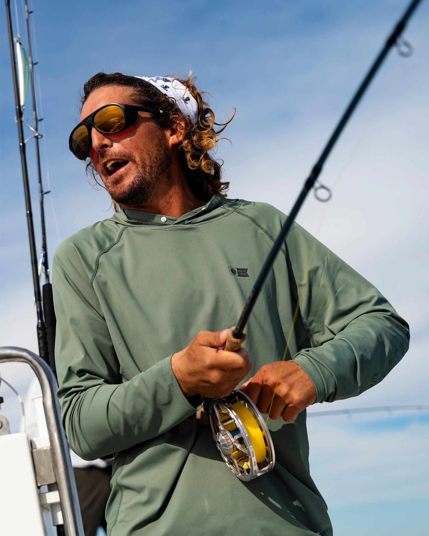 Sporting the Salty Crew Apex Perforated Sunshirt - Agave, which delivers UPF 50+ sun protection in a breathable design, a person wearing sunglasses and a bandana fishes on a boat with rod and reel beneath a blue sky.