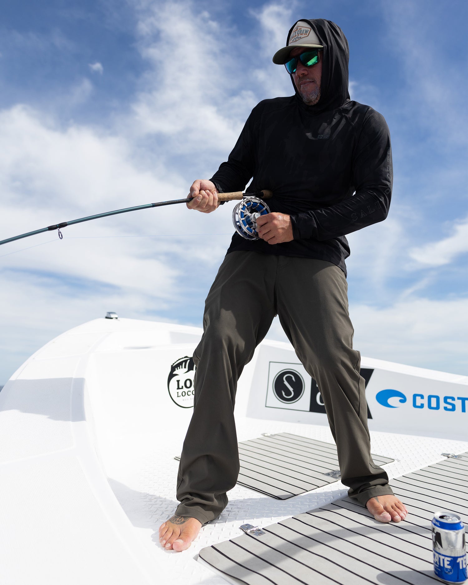 Wearing Salty Crew’s Apex Perforated Sunshirt - Ranger, a person in sunglasses and a hood stands barefoot on a boat, holding a fishing rod with a can resting on the deck and blue sky overhead.