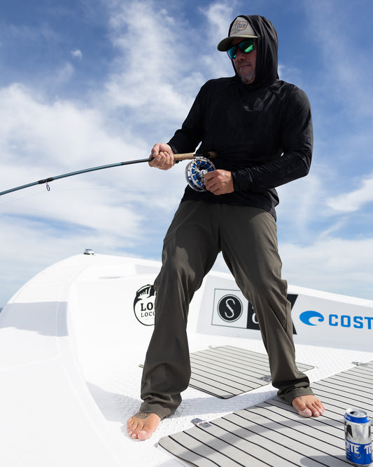 Wearing Salty Crew’s Apex Perforated Sunshirt - Ranger, a person in sunglasses and a hood stands barefoot on a boat, holding a fishing rod with a can resting on the deck and blue sky overhead.