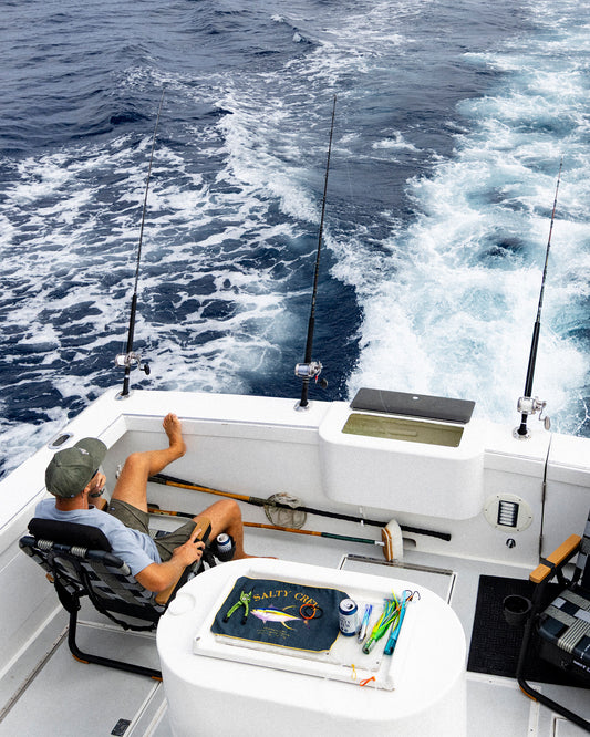 A person sits in a Parkit x Salty Crew Eclipse Recliner on the back deck of a boat, with three fishing rods set up against the transom while the vessel leaves a wake in the water.