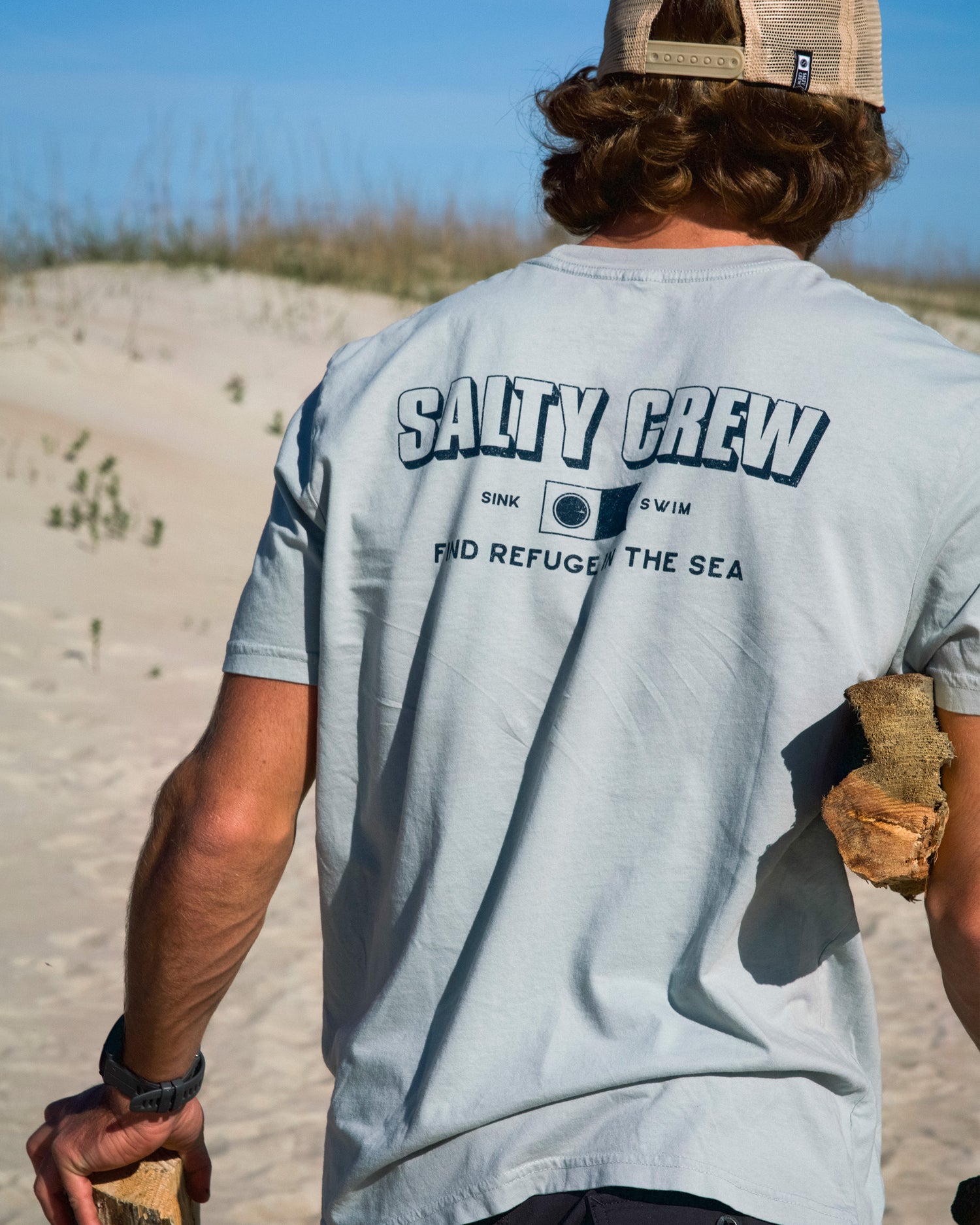A man stands on a sandy beach holding driftwood, wearing the Salty Crew Simple Original Tee in Aqua Grey and a tan cap. Crafted from 100% cotton soft spun fabric, this comfortable T-shirt features screenprinted graphics with the phrase “Sink or Swim. Find Refuge in the Sea.” The sky and sand form the backdrop to this stylish look.