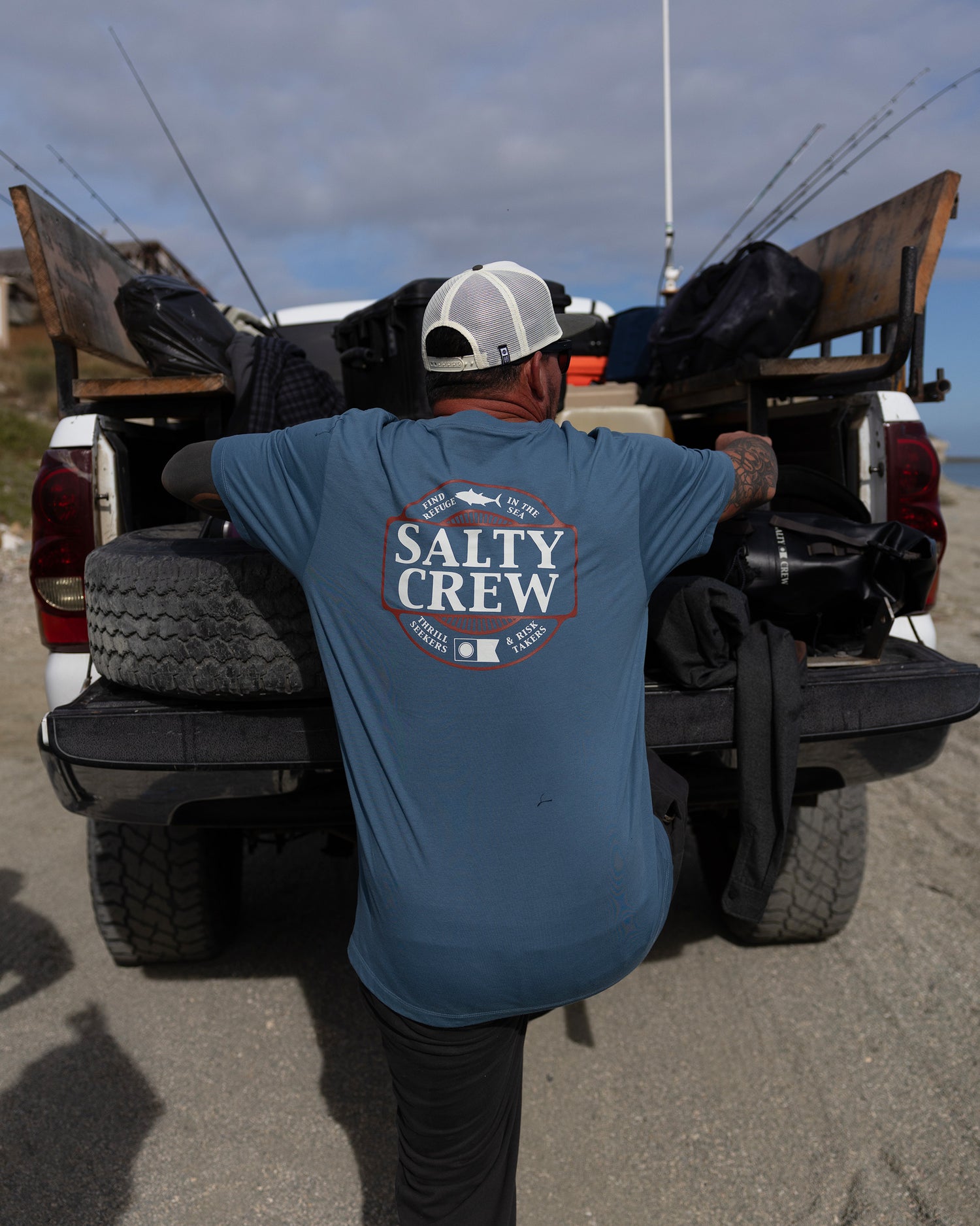 Wearing the Salty Crew Split Up Crew Tee in Slate, a person stands at the back of a truck with fishing gear and rods, getting ready for a day outdoors on a sandy beach.