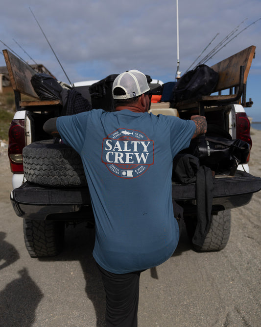 Wearing the Salty Crew Split Up Crew Tee in Slate, a person stands at the back of a truck with fishing gear and rods, getting ready for a day outdoors on a sandy beach.