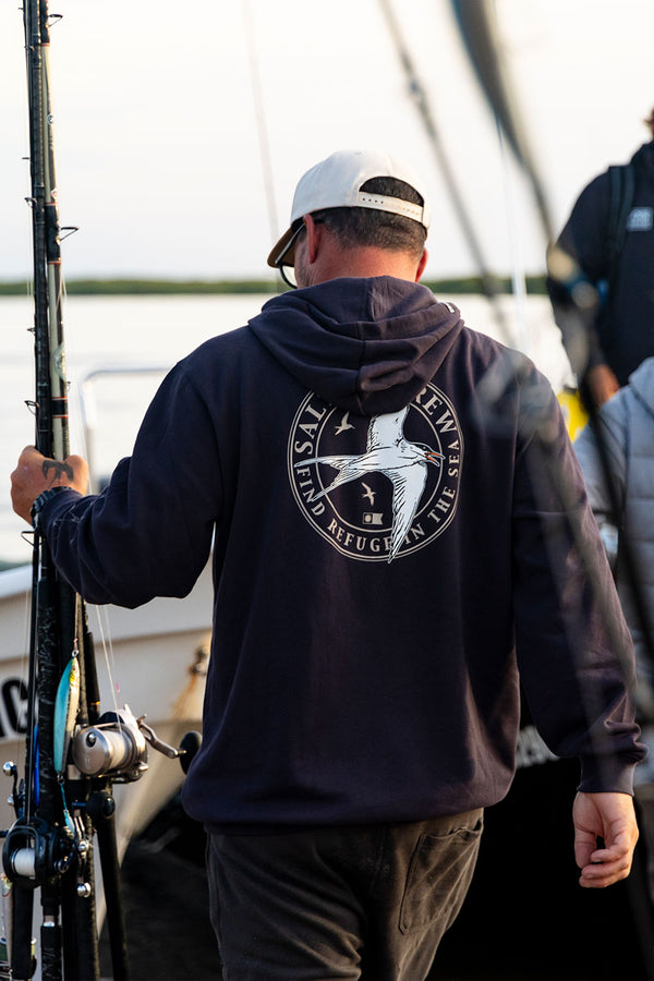 A man in the Salty Crew Tern Fleece - Dark Grey hoodie, featuring a circular graphic and text, stands on a dock holding a fishing rod, with boats and another person visible in the background.