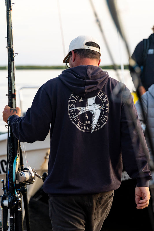 A man in the Salty Crew Tern Fleece - Dark Grey hoodie, featuring a circular graphic and text, stands on a dock holding a fishing rod, with boats and another person visible in the background.