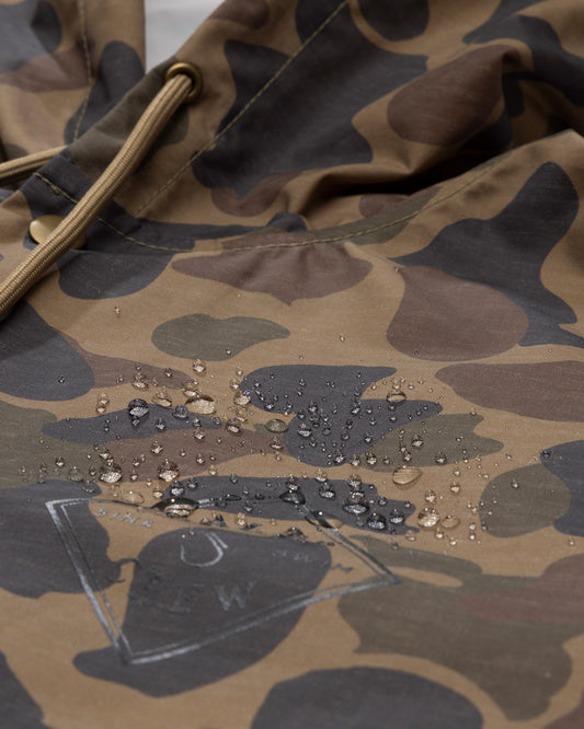 Close-up of the Salty Crew Tippet Snap Jacket in Sand Camo, highlighting water droplets beading on its camouflage-patterned heavyweight nylon shell, which features a 5K waterproof rating. Drawstrings and a subtle white screenprinted logo are also visible.