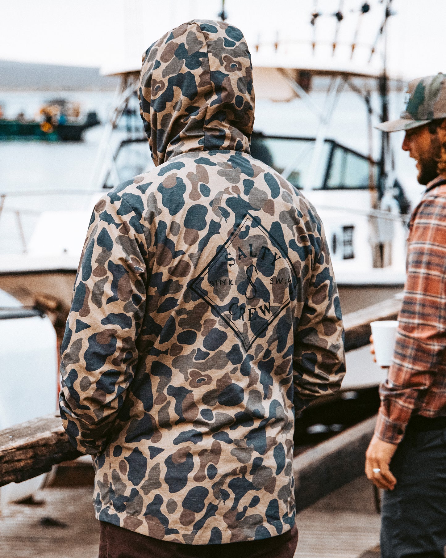 Two people stand near a docked boat. One, facing away, wears the Salty Crew Tippet Snap Jacket in Sand Camo—a heavyweight nylon jacket with a hood and branded text on the back. The other, in a cap and plaid shirt, holds a cup. Water and boats fill the background.