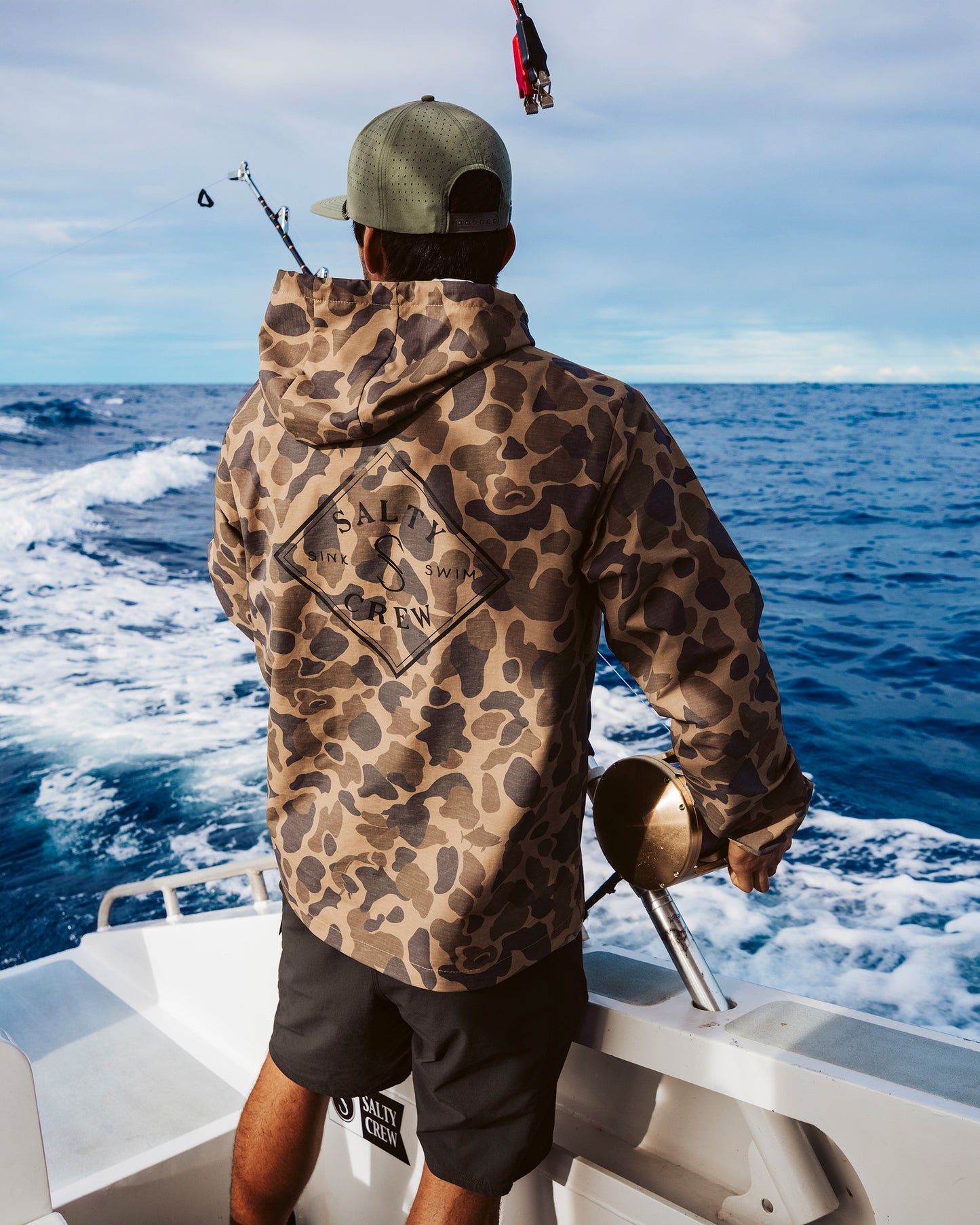 Wearing the Salty Crew Tippet Snap Jacket in Sand Camo and a cap, a person stands on a boat with a fishing rod, gazing out over the blue ocean beneath a partly cloudy sky.
