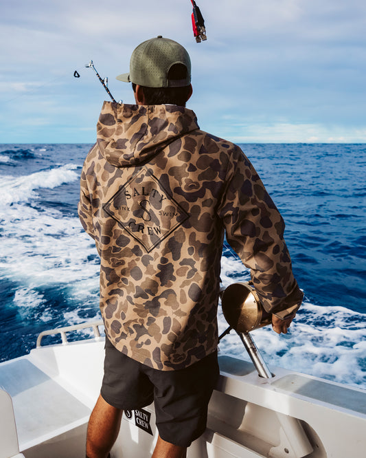 Wearing the Salty Crew Tippet Snap Jacket in Sand Camo and a cap, a person stands on a boat with a fishing rod, gazing out over the blue ocean beneath a partly cloudy sky.