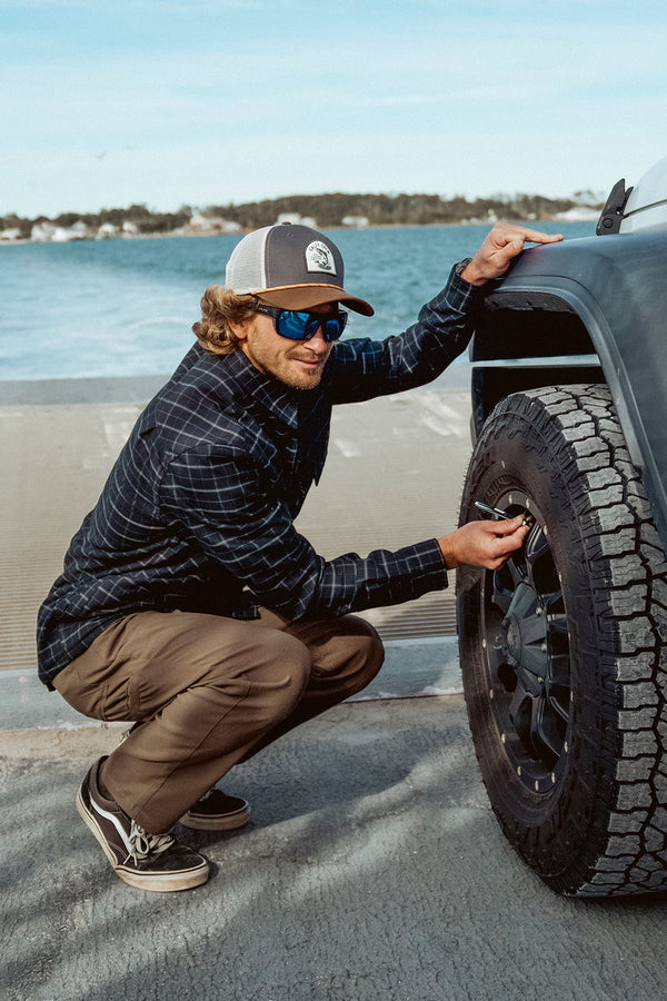 A man wearing sunglasses and a cap squats beside a black truck near a waterfront on a sunny day, checking the tire pressure with a gauge, dressed in Salty Crew’s Transom Tech Pant in Washed Brown—quick-dry clothing made from recycled performance fabric.