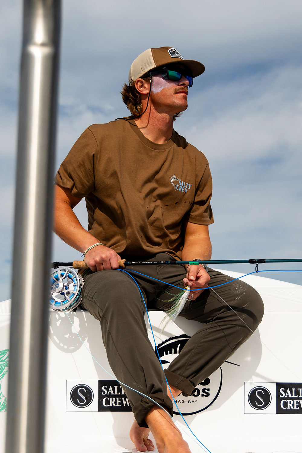 A person wearing sunglasses, a Salty Crew Transom Tech Pant in Washed Brown made from quick-dry, recycled performance fabric, and a cap is sitting on a boat holding fishing gear with blue fishing line. The background features a cloudy sky, with boat logos visible below them.