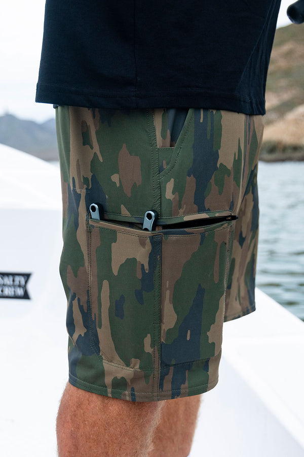 A person wearing Salty Crew Transom 19″ Tech Short - Ranger, featuring cargo pockets including a large zippered pocket holding two blue-handled tools, stands on a boat with water and hills in the background.