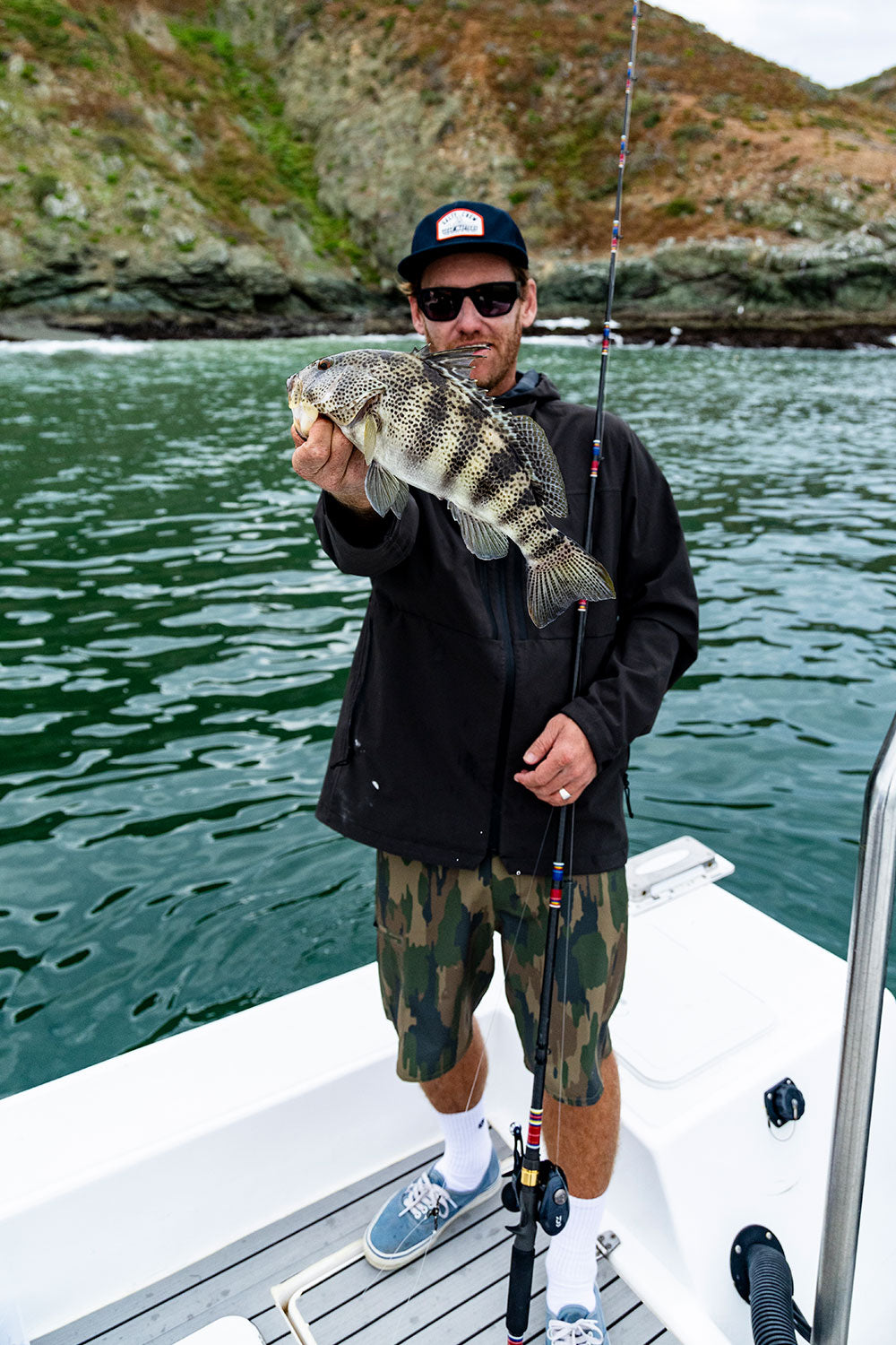 A person in sunglasses and a cap stands on a boat, wearing Salty Crew’s Transom 19″ Tech Short - Ranger while holding a freshly caught fish in one hand and a fishing rod in the other, with water and rocky hills in the background.